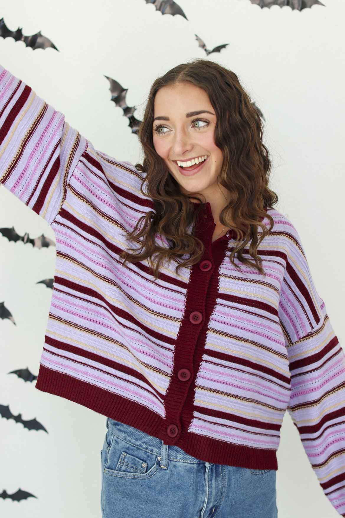 A woman wearing a knit cardigan with horizontal stripes in dark purple, light pink, and white, paired with blue denim skirt, posing against a white wall decorated with flying bat cutouts.