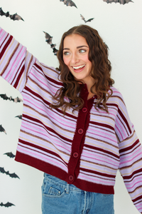 A woman wearing a knit cardigan with horizontal stripes in dark purple, light pink, and white, paired with blue denim skirt, posing against a white wall decorated with flying bat cutouts.
