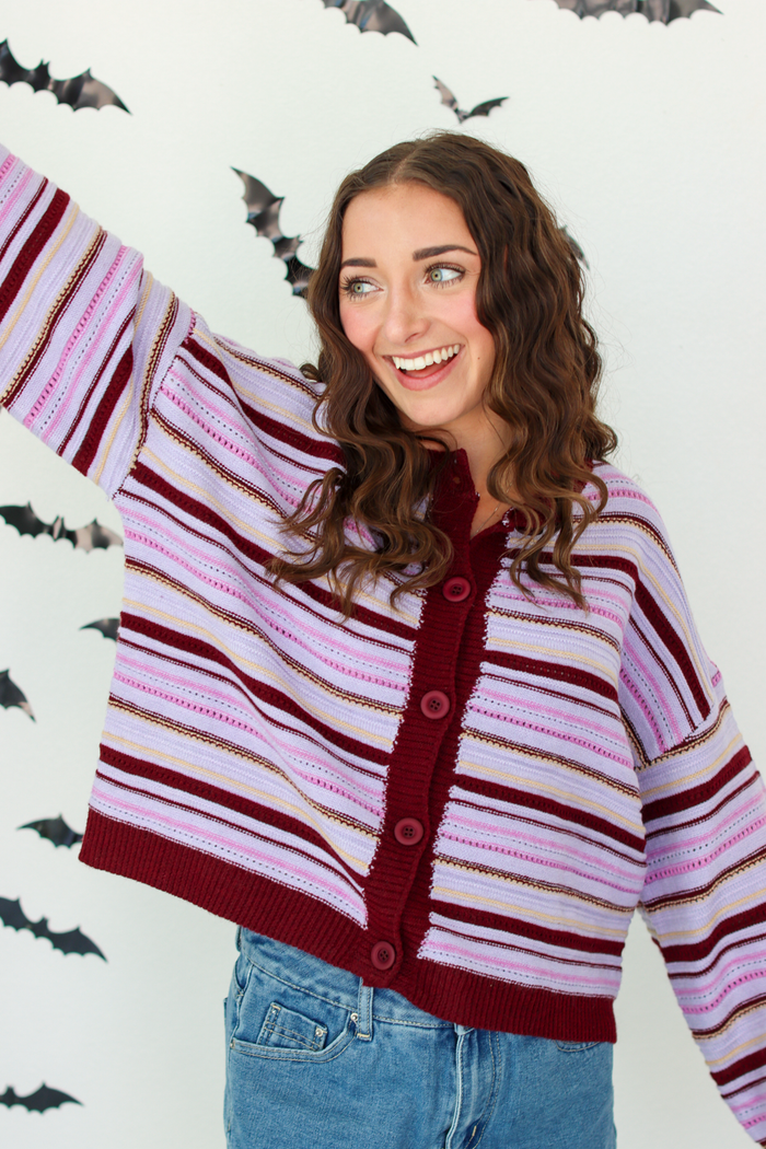 A woman wearing a knit cardigan with horizontal stripes in dark purple, light pink, and white, paired with blue denim skirt, posing against a white wall decorated with flying bat cutouts.