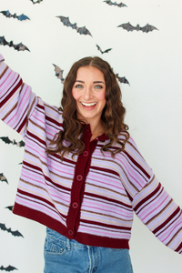 A woman with long brown hair smiling and raising her right arm, wearing a knit cardigan with horizontal stripes in dark purple, light pink, and white, paired with blue denim skirt. The background is a white wall decorated with black bat silhouettes.
