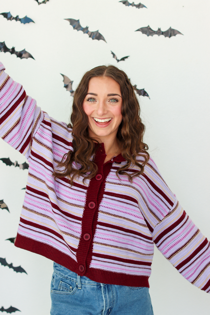 A woman with long brown hair smiling and raising her right arm, wearing a knit cardigan with horizontal stripes in dark purple, light pink, and white, paired with blue denim skirt. The background is a white wall decorated with black bat silhouettes.
