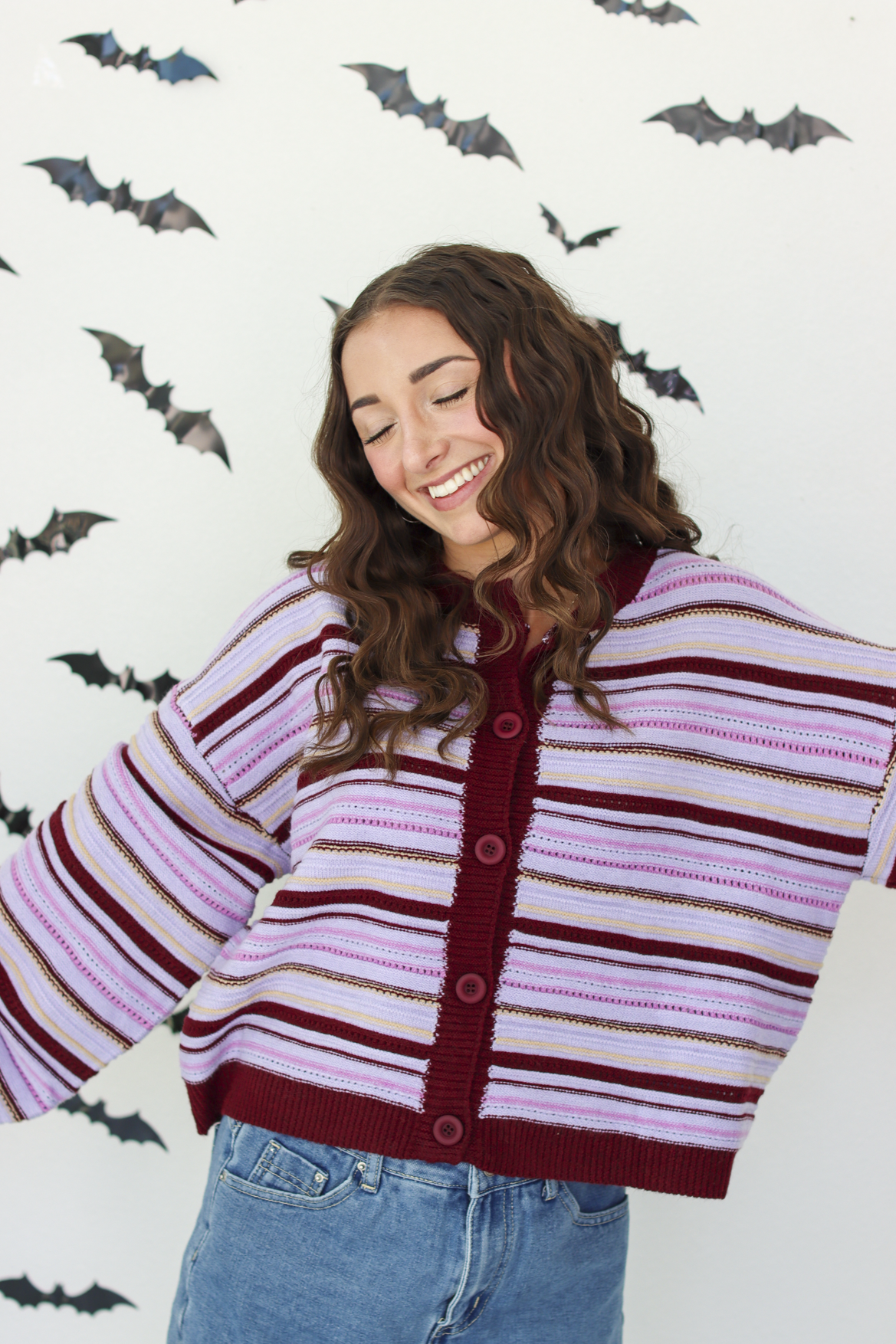 A woman wearing a knit cardigan with horizontal stripes in dark purple, light pink, and white, paired with blue denim skirt, posing against a white wall decorated with flying bat cutouts.