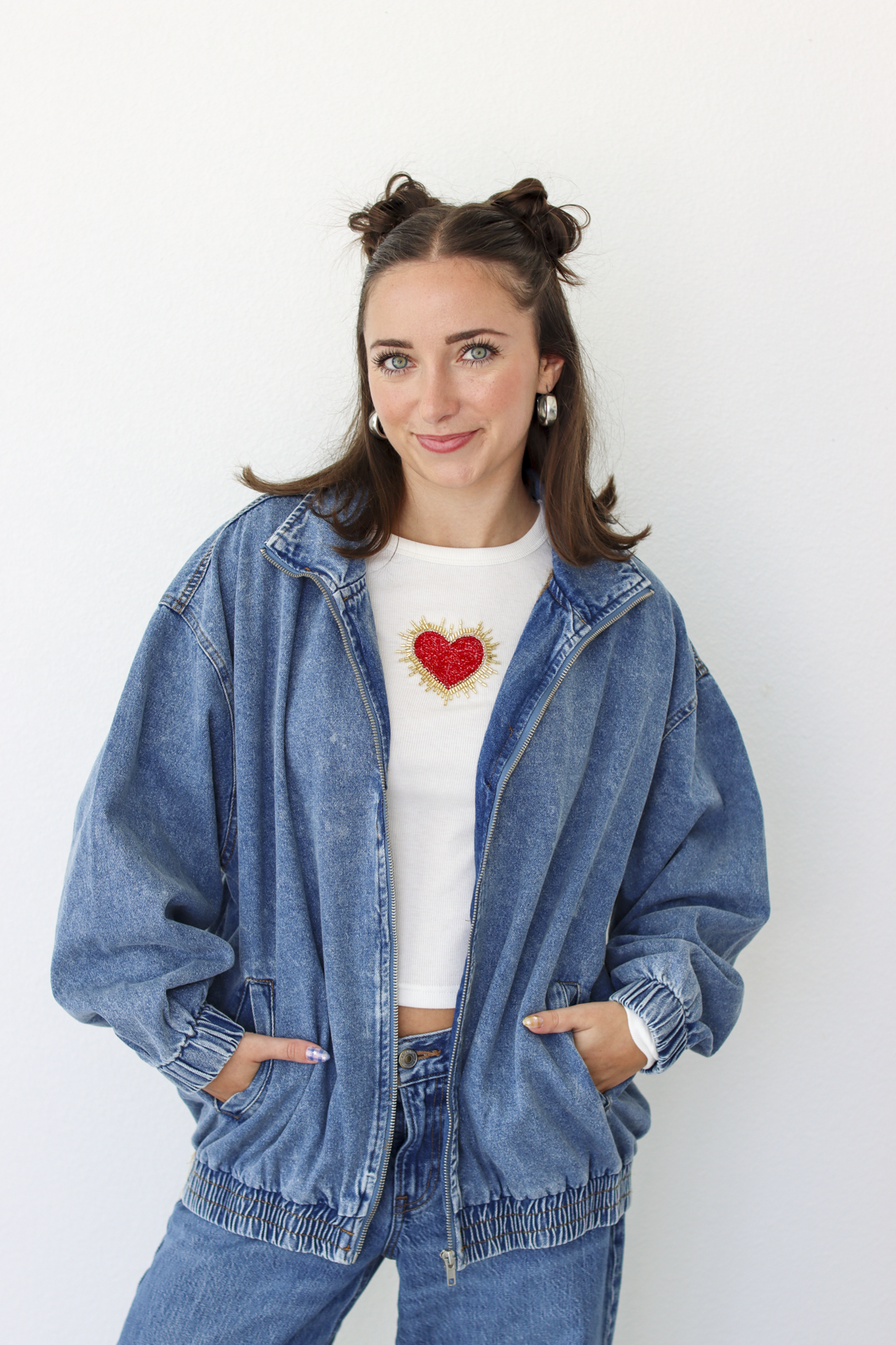 A woman wearing a blue denim zip-up jacket over a white cropped t-shirt with a red heart graphic, paired with blue jeans, standing against a plain white wall.