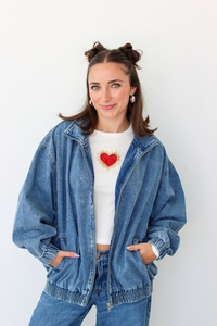 A woman wearing a blue denim zip-up jacket over a white cropped t-shirt with a red heart graphic, paired with blue jeans, standing against a plain white wall.