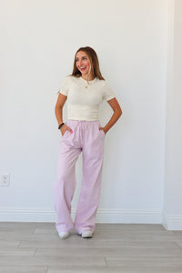 Woman wearing a white top and light pink pants standing against a white wall.