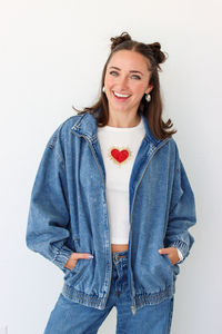 A smiling woman with her hair in two buns wears an oversized light-wash blue denim jacket over a white crop top with a red heart graphic, paired with blue jeans, against a plain white background.