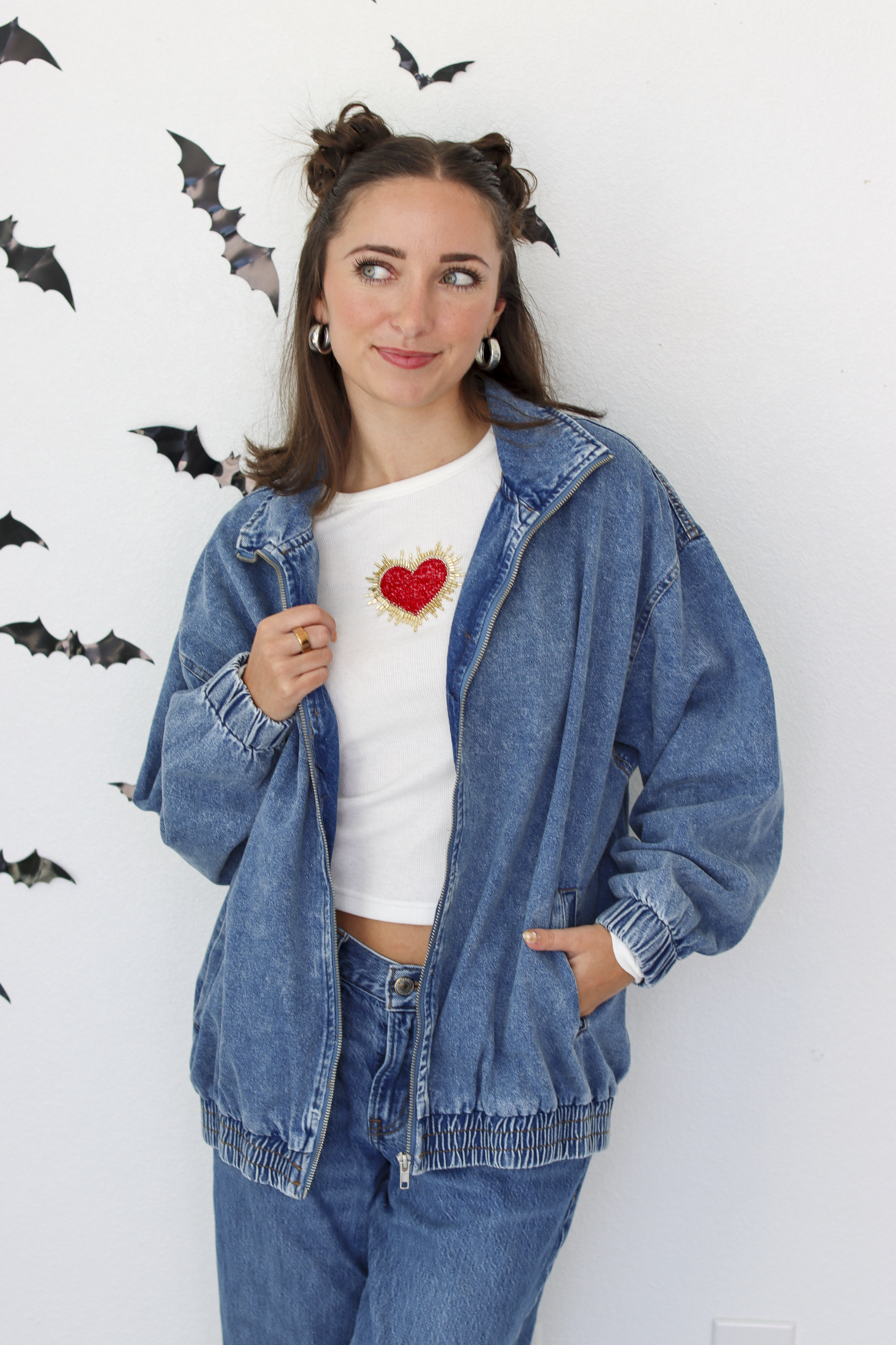 A woman wearing a blue denim zip-up hoodie over a white t-shirt with a red heart graphic, paired with matching blue jeans. She is standing against a white wall decorated with black bat cutouts.