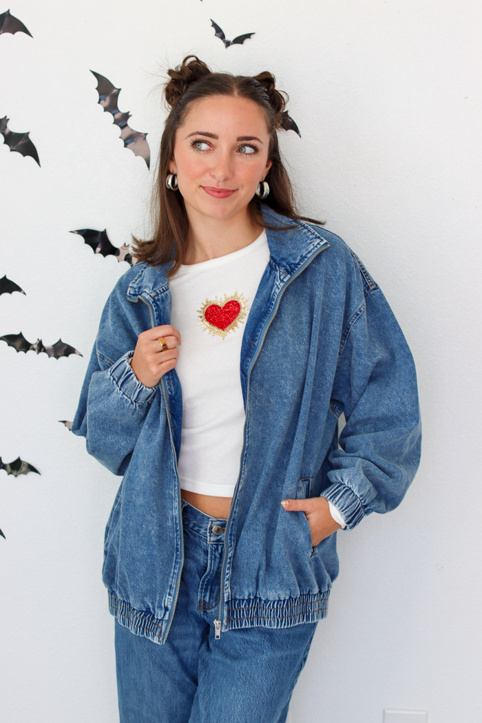A woman wearing a blue denim zip-up hoodie over a white t-shirt with a red heart graphic, paired with matching blue jeans. She is standing against a white wall decorated with black bat cutouts.