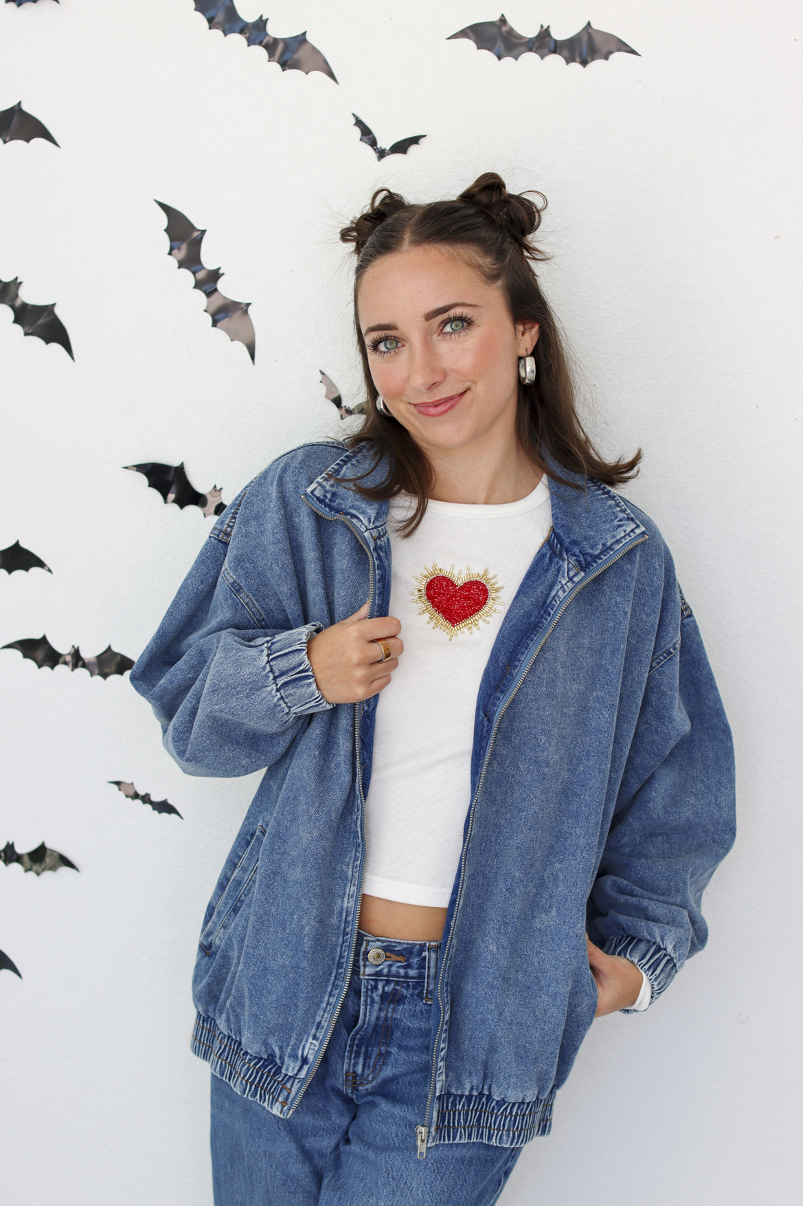 A woman posing in a blue denim jacket over a white crop top with a red heart graphic, paired with blue jeans, set against a white wall decorated with bat silhouettes.