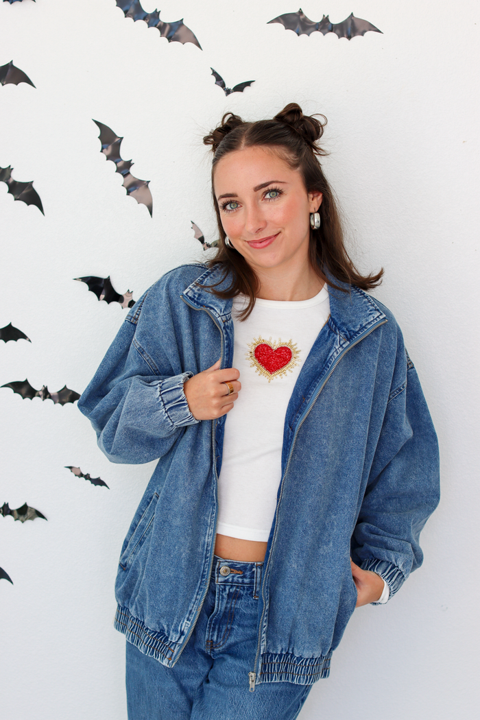 A woman posing in a blue denim jacket over a white crop top with a red heart graphic, paired with blue jeans, set against a white wall decorated with bat silhouettes.