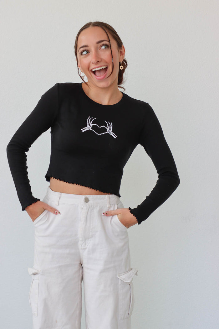 girl wearing black long sleeved cropped top with embroidered white skeleton hands in a heart