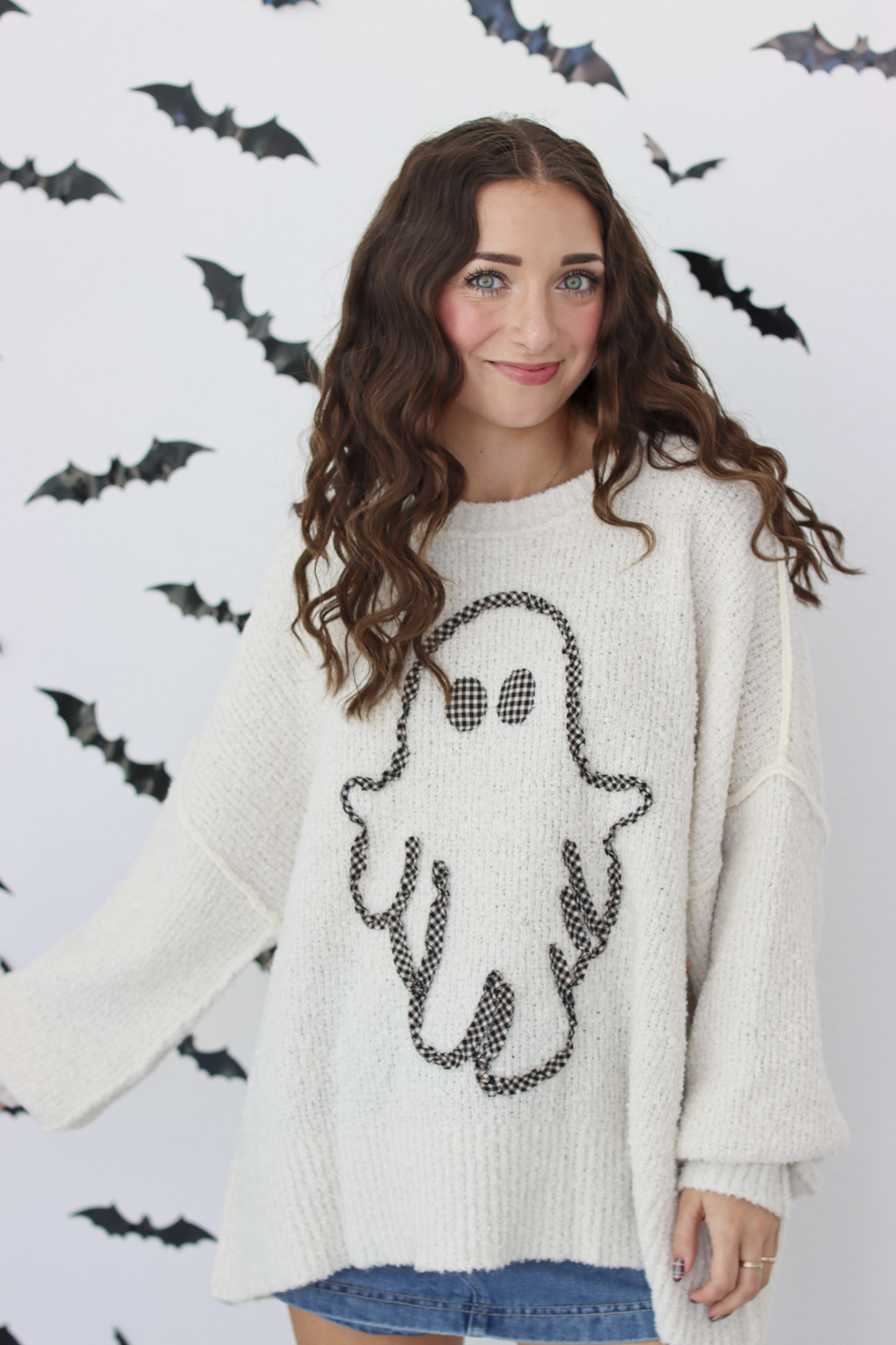 A woman wearing a cream-colored knit sweater with a black ghost graphic on the front, posing against a white wall decorated with black bat silhouettes.