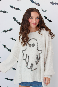 A woman wearing a cream-colored knit sweater with a black ghost graphic on the front, posing against a white wall decorated with black bat silhouettes.