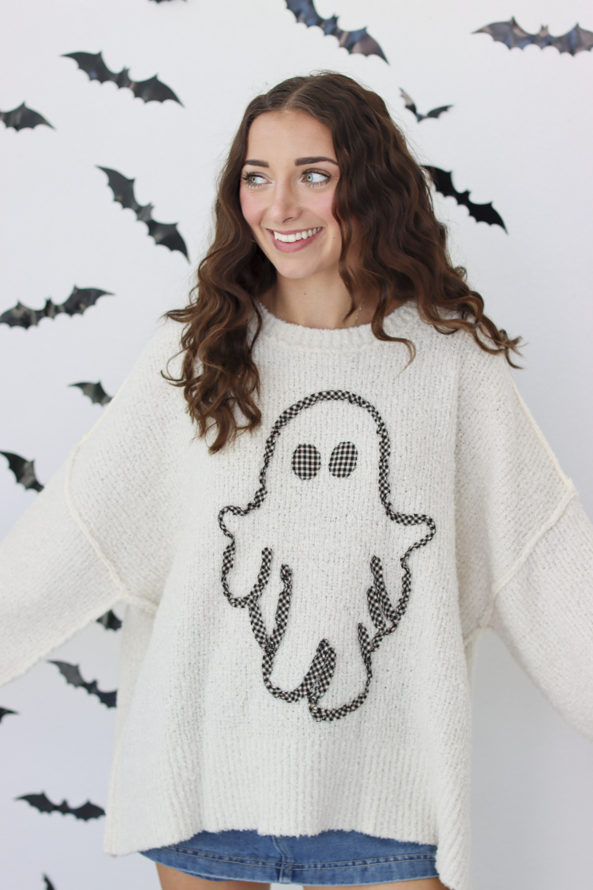 A woman wearing a cream-colored knit sweater with a black ghost graphic on the front, posing against a white wall decorated with black bat silhouettes.