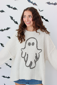 A woman wearing a cream-colored knit sweater with a black ghost graphic on the front, posing against a white wall decorated with black bat silhouettes.