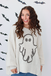 A woman wearing a cream-colored knit sweater with a black ghost graphic on the front, posing against a white wall decorated with black bat silhouettes.
