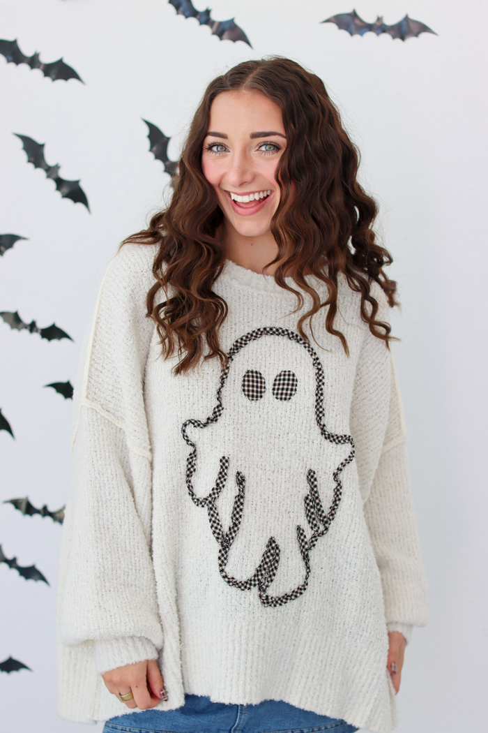 A woman wearing a cream-colored knit sweater with a black ghost graphic on the front, posing against a white wall decorated with black bat silhouettes.