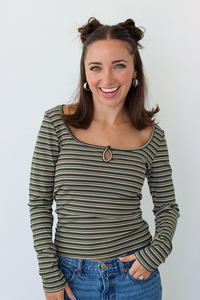 A woman with her hair in two buns is smiling and wearing a long-sleeve, scoop-neck top with thin horizontal stripes in olive green, black, and beige. She is also wearing blue jeans and silver hoop earrings, standing against a plain white background.