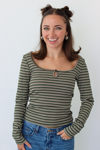 A smiling woman with brown hair in two buns wears a long-sleeve scoop-neck top with thin horizontal olive green, black, and beige stripes, paired with blue jeans, against a plain white background.