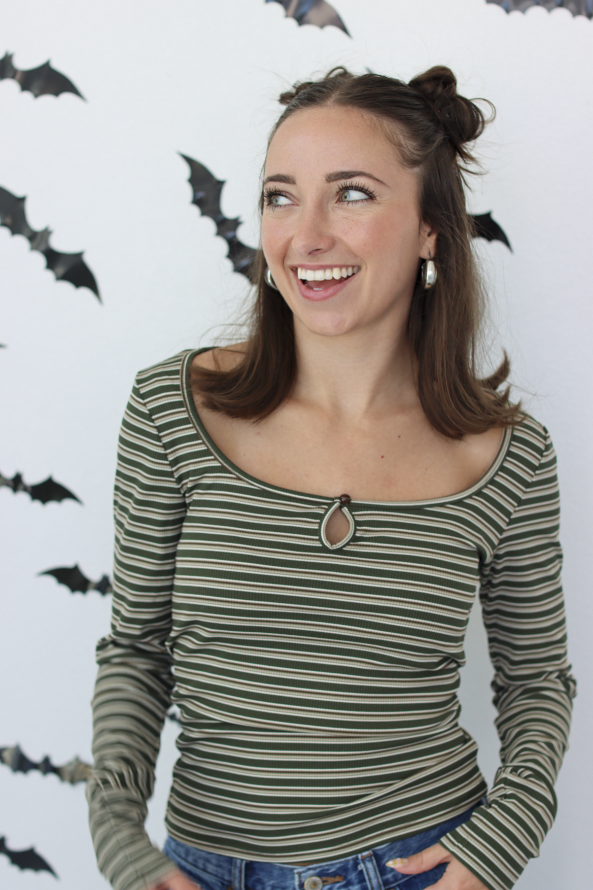 A woman smiling and looking to the side, wearing a long-sleeve knit top with horizontal olive green and black stripes and a keyhole cutout at the neckline. She is standing against a white wall decorated with black bat cutouts.