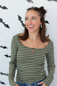 A woman smiling and looking to the side, wearing a long-sleeve knit top with horizontal olive green and black stripes and a keyhole cutout at the neckline. She is standing against a white wall decorated with black bat cutouts.