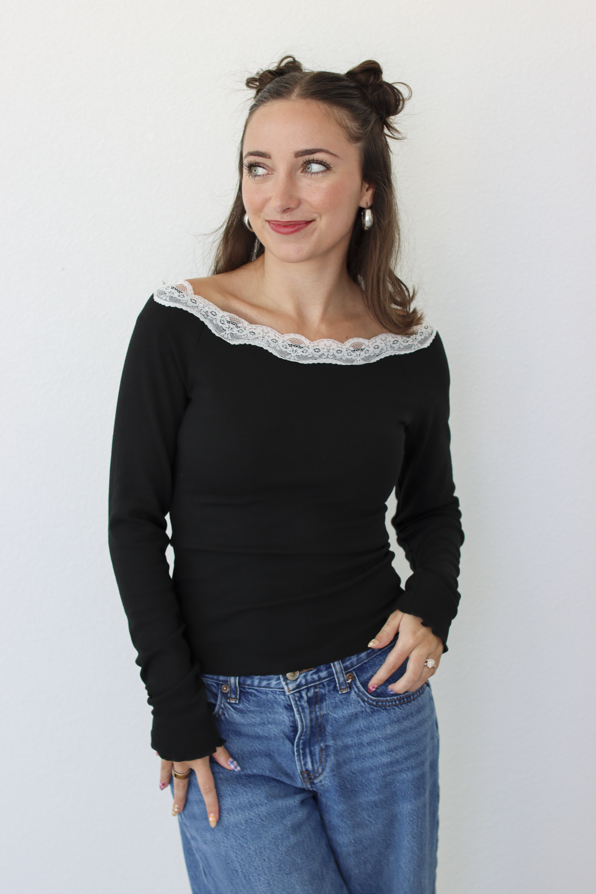A woman with her hair in two buns is wearing a black off-the-shoulder long-sleeve top with a white lace trim and blue jeans, standing against a plain white wall.