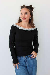 A woman with her hair in two buns is wearing a black off-the-shoulder long-sleeve top with a white lace trim and blue jeans, standing against a plain white wall.