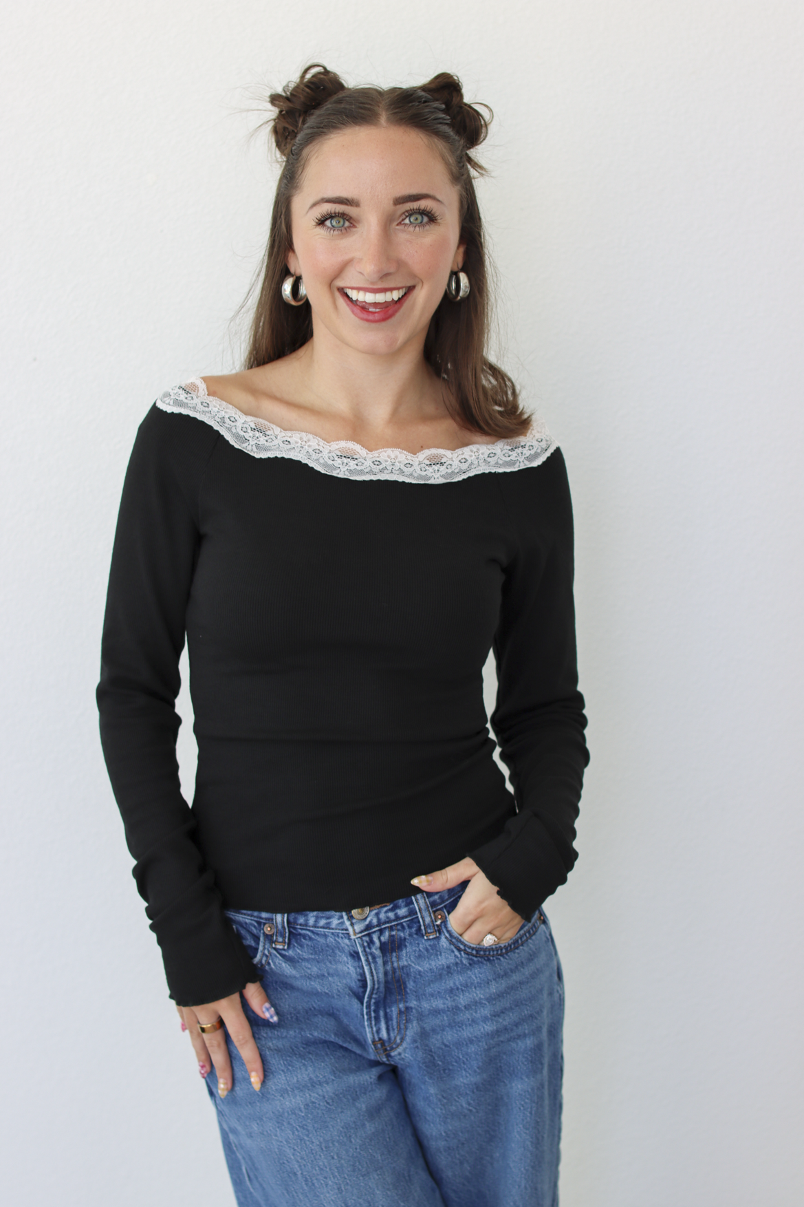 A woman with her hair in two buns is wearing a black off-the-shoulder long-sleeve top with a white lace trim and blue jeans, standing against a plain white wall.