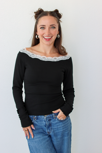 A woman with her hair in two buns is wearing a black off-the-shoulder long-sleeve top with a white lace trim and blue jeans, standing against a plain white wall.