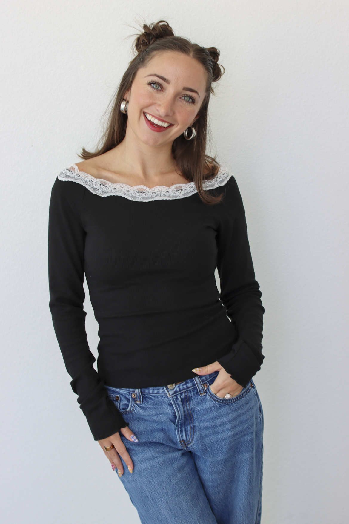 A woman with her hair in two buns is wearing a black off-the-shoulder long-sleeve top with a white lace trim and blue jeans, standing against a plain white wall.