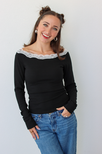 A woman with her hair in two buns is wearing a black off-the-shoulder long-sleeve top with a white lace trim and blue jeans, standing against a plain white wall.