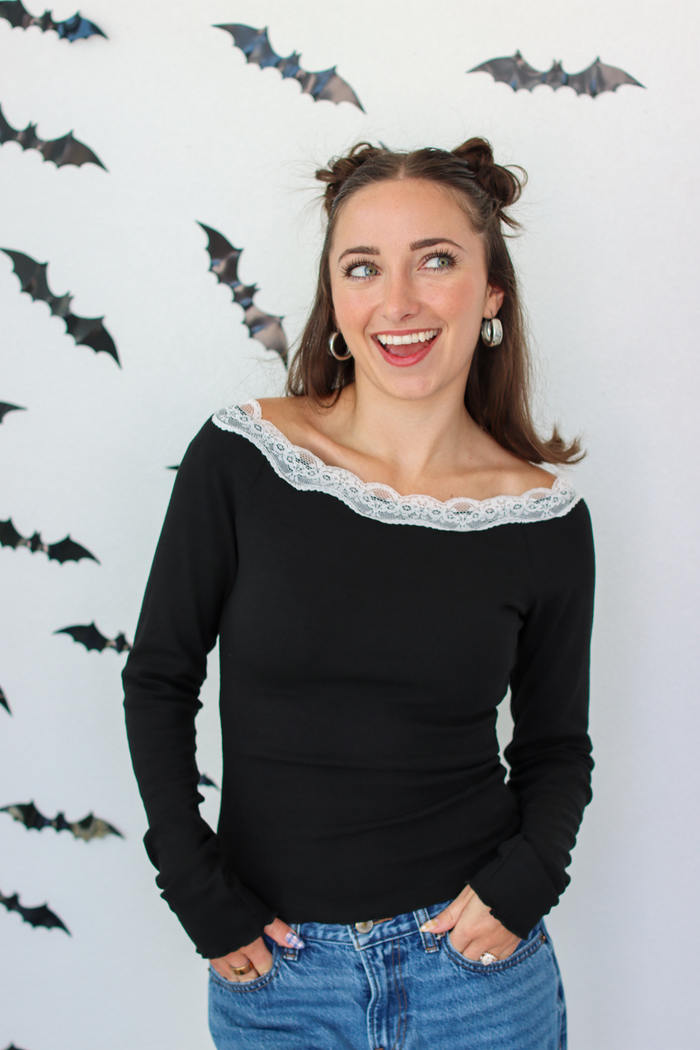 A woman with her hair in two buns wears a black long-sleeve top with a white lace off-the-shoulder collar, paired with blue jeans, standing against a white wall decorated with small bat cutouts.