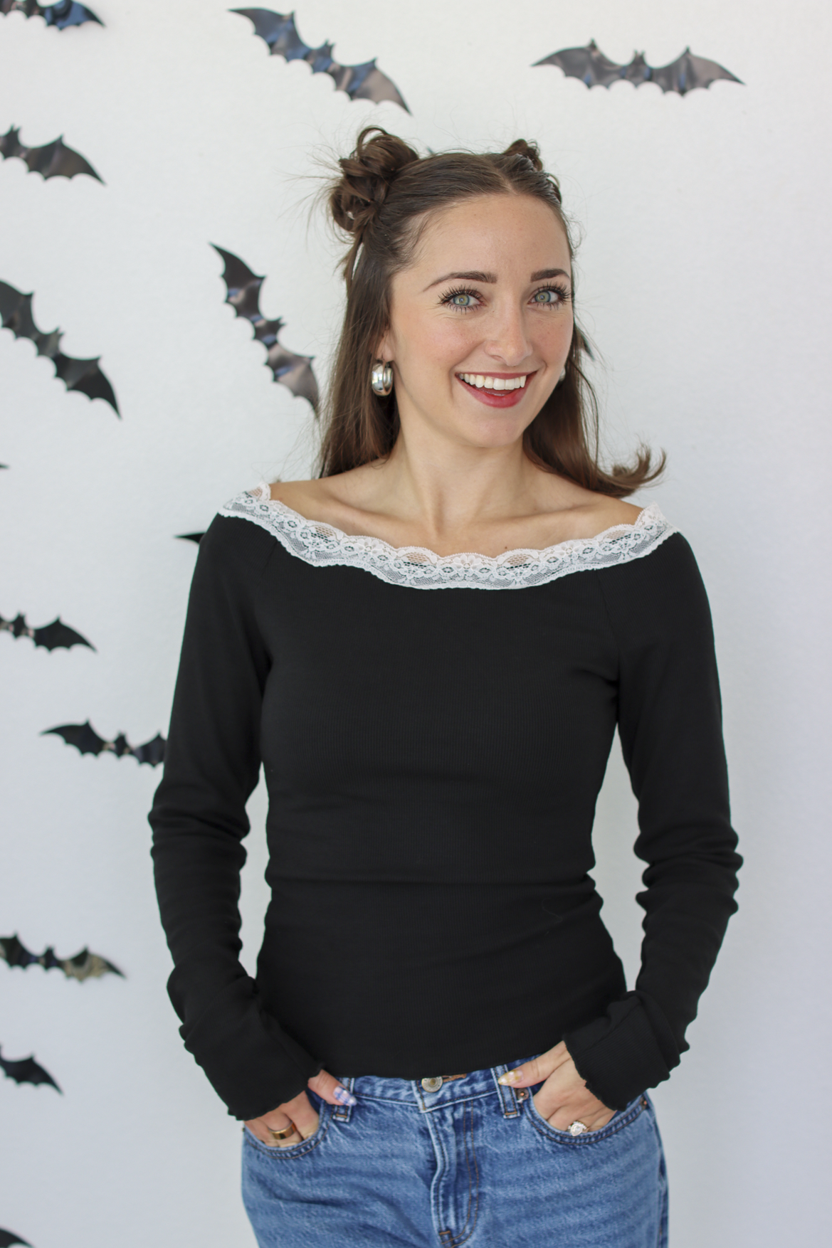 A woman with her hair in two buns wears a black long-sleeve top with a white lace off-the-shoulder collar, paired with blue jeans, standing against a white wall decorated with small bat cutouts.