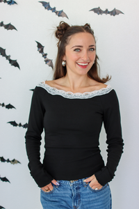 A woman with her hair in two buns wears a black long-sleeve top with a white lace off-the-shoulder collar, paired with blue jeans, standing against a white wall decorated with small bat cutouts.