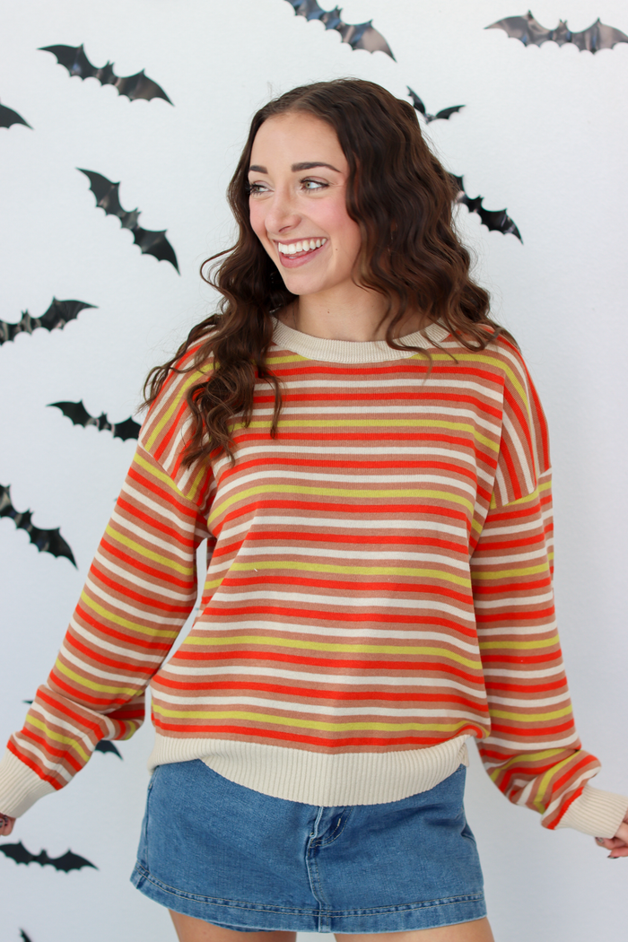A woman wearing a long-sleeve striped knit sweater with horizontal bands of orange, yellow, red, and cream, paired with a blue denim skirt, standing against a white wall decorated with black bat silhouettes.