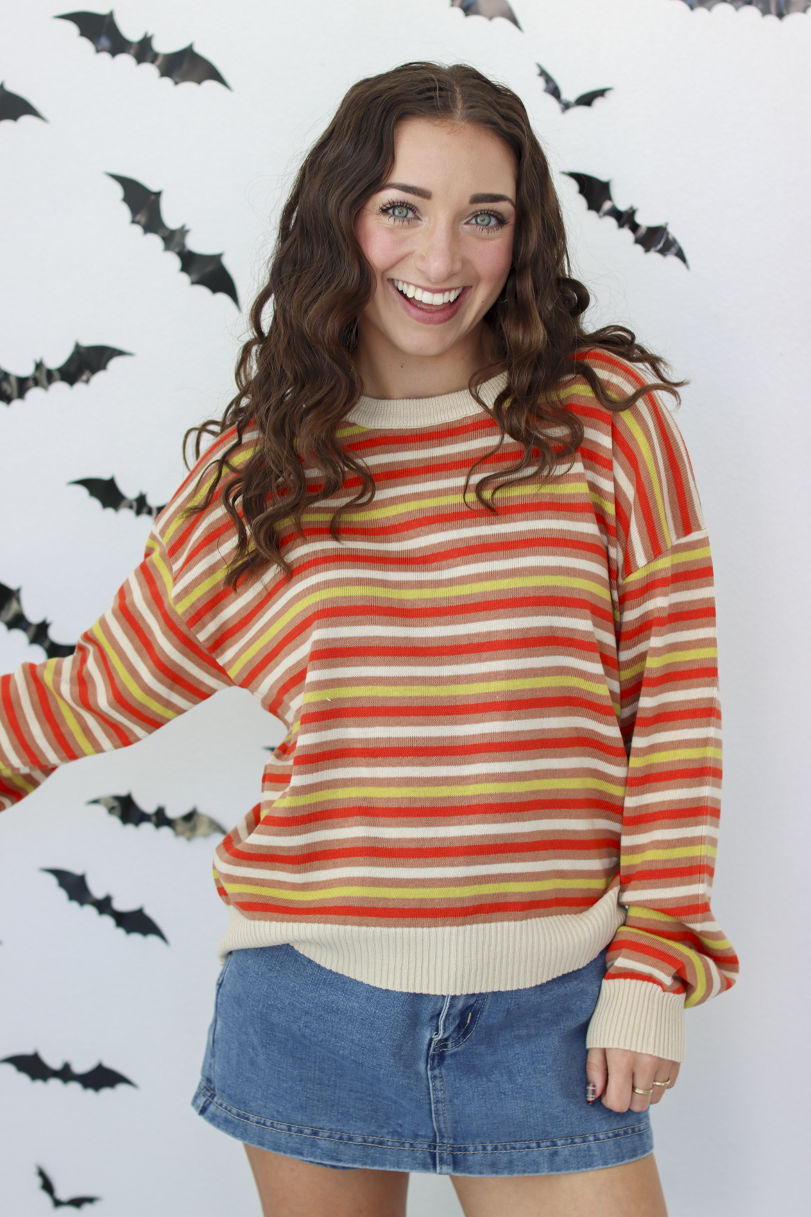 A woman wearing a long-sleeve striped knit sweater with horizontal bands of orange, yellow, red, and cream, paired with a blue denim skirt, standing against a white wall decorated with black bat silhouettes.