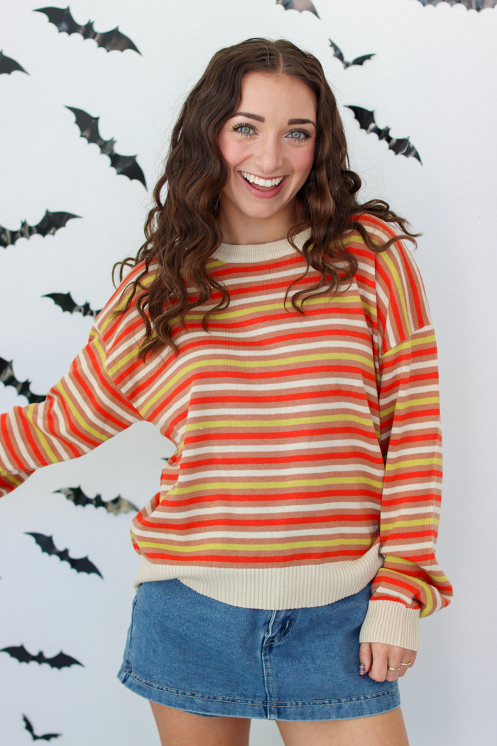 A woman wearing a long-sleeve striped knit sweater with horizontal bands of orange, yellow, red, and cream, paired with a blue denim skirt, standing against a white wall decorated with black bat silhouettes.