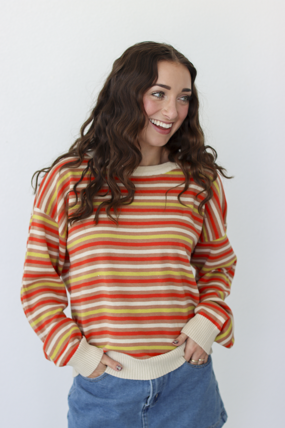 A woman with long wavy brown hair smiling and looking to the side, wearing a long-sleeve crewneck sweater with horizontal stripes in orange, yellow, and cream, paired with blue denim skirt against a plain white background.