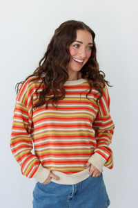 A woman with long wavy brown hair smiling and looking to the side, wearing a long-sleeve crewneck sweater with horizontal stripes in orange, yellow, and cream, paired with blue denim skirt against a plain white background.