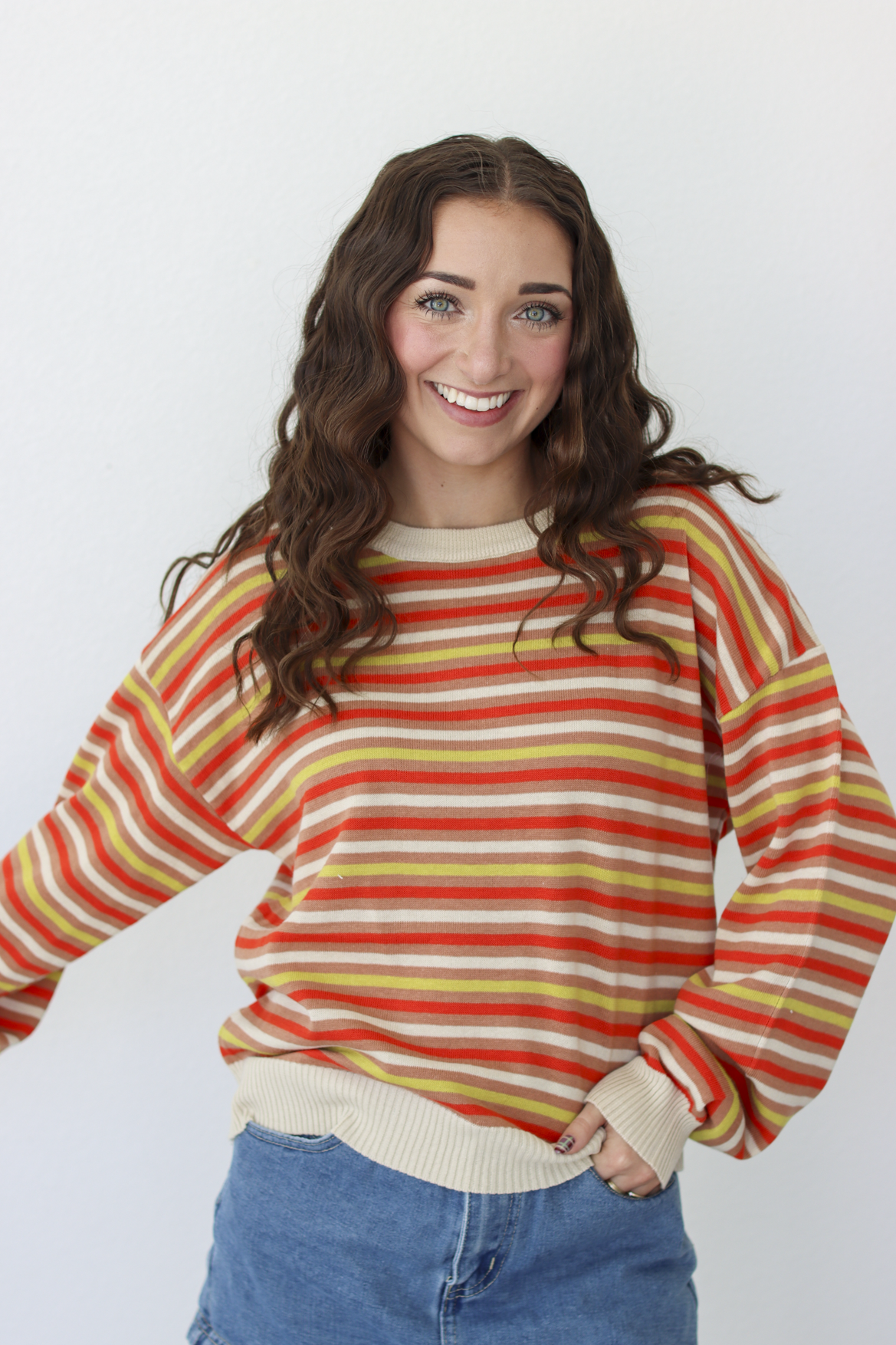 A smiling young woman with long wavy brown hair wears a long-sleeve crewneck sweater with horizontal stripes in orange, yellow, and cream, paired with blue denim skirt against a plain white background.