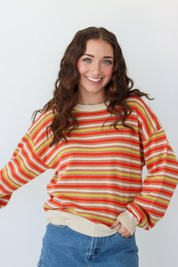 A smiling young woman with long wavy brown hair wears a long-sleeve crewneck sweater with horizontal stripes in orange, yellow, and cream, paired with blue denim skirt against a plain white background.