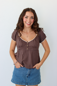 A smiling woman with long brown hair stands against a plain white wall wearing a brown gingham short-sleeve top with white trim and a blue denim mini skirt.