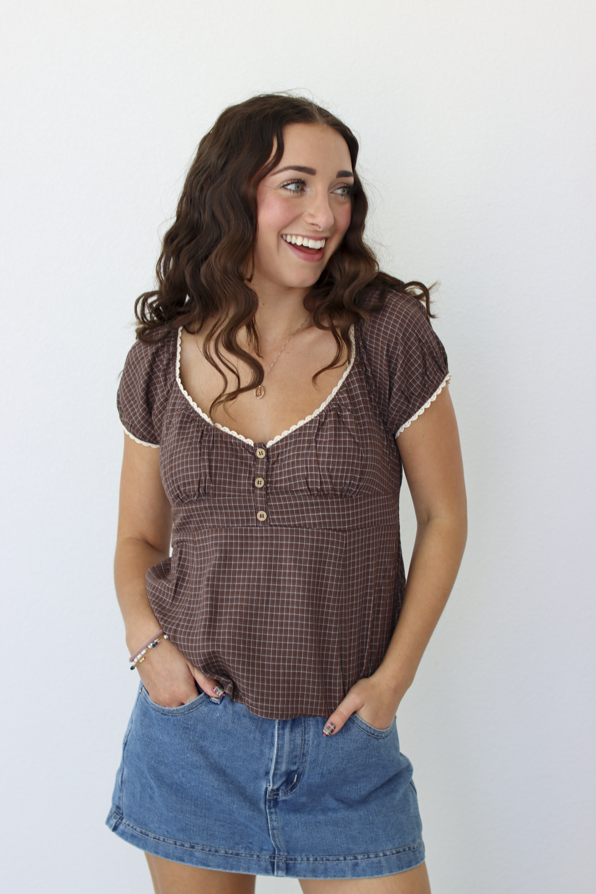 A smiling woman with long brown hair stands against a plain white wall wearing a brown gingham short-sleeve top with white trim and a blue denim mini skirt.