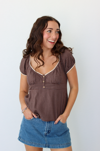 A smiling woman with long brown hair stands against a plain white wall wearing a brown gingham short-sleeve top with white trim and a blue denim mini skirt.