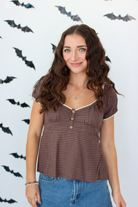 A woman with long wavy brown hair wearing a brown checkered short-sleeve top with white lace trim and blue denim skirt, standing against a white wall decorated with black bat cutouts.