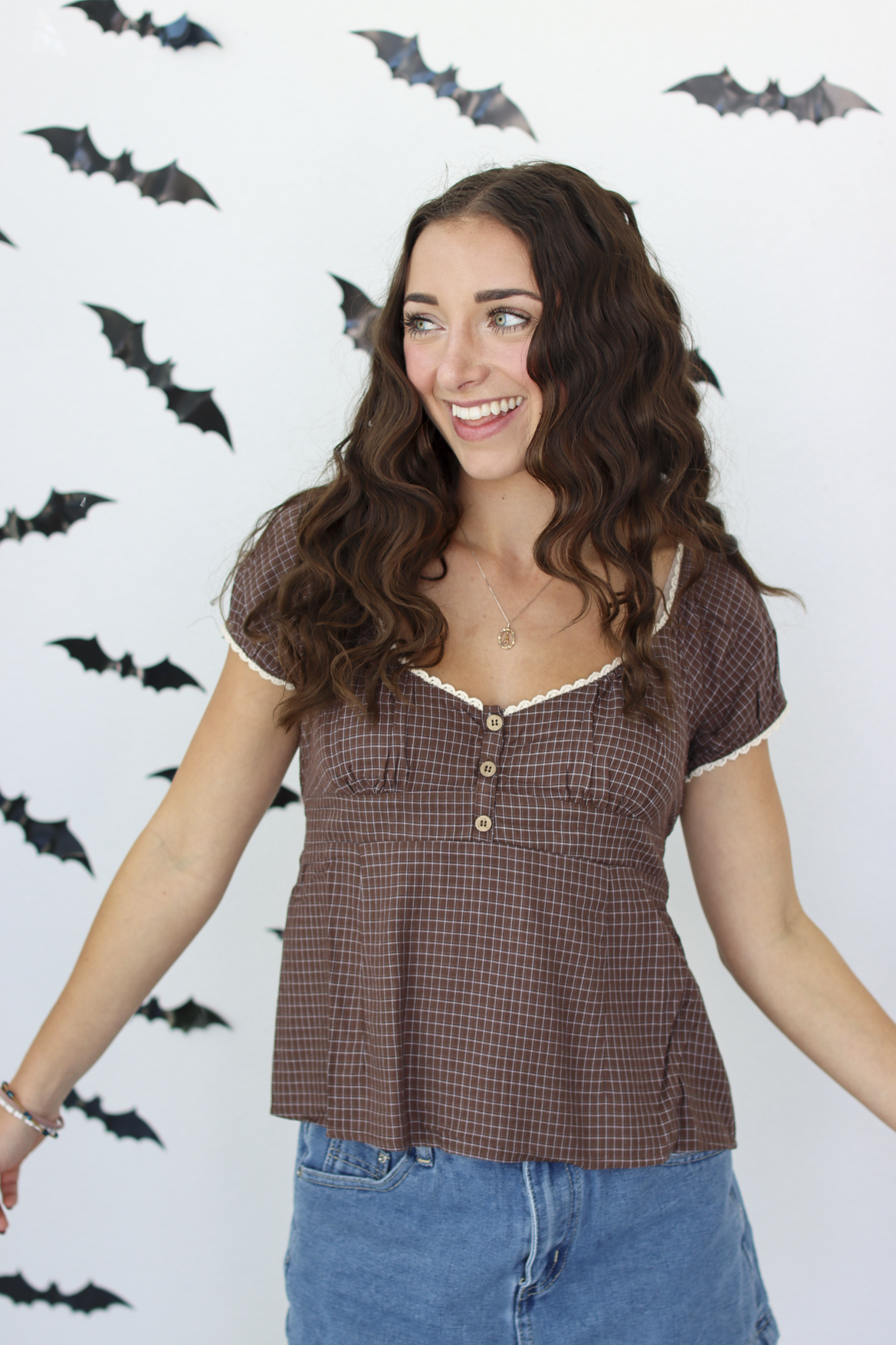 A woman with long wavy brown hair wearing a brown checkered short-sleeve top with white lace trim and blue denim skirt, standing against a white wall decorated with black bat cutouts.