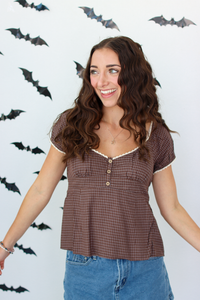 A woman with long wavy brown hair wearing a brown checkered short-sleeve top with white lace trim and blue denim skirt, standing against a white wall decorated with black bat cutouts.