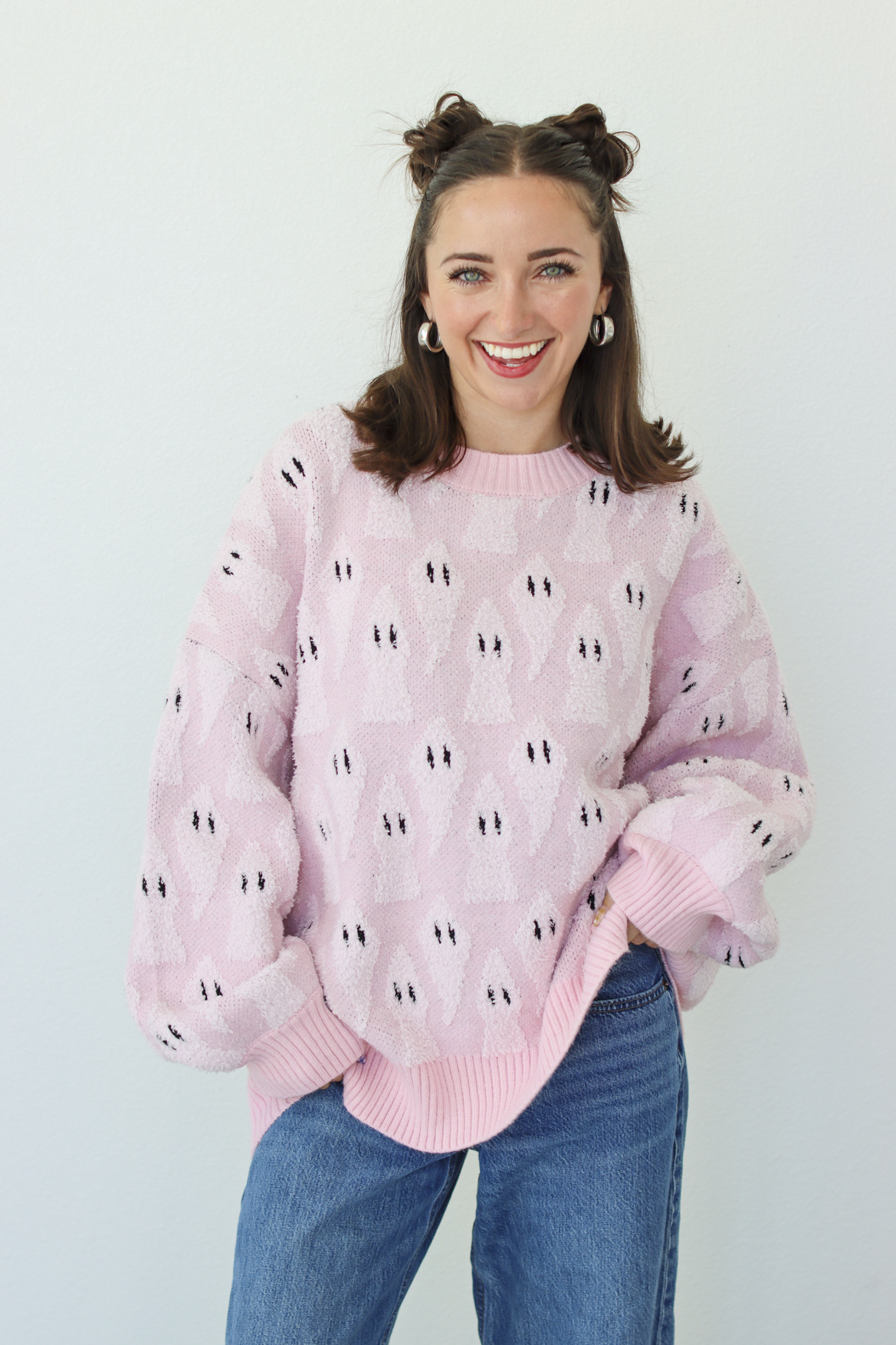 A smiling woman with her hair in two buns wears a light pink fuzzy knit sweater with a repeating black and white ghost pattern, paired with blue jeans, against a plain light gray background.