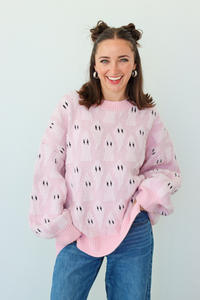A smiling woman with her hair in two buns wears a light pink fuzzy knit sweater with a repeating black and white ghost pattern, paired with blue jeans, against a plain light gray background.