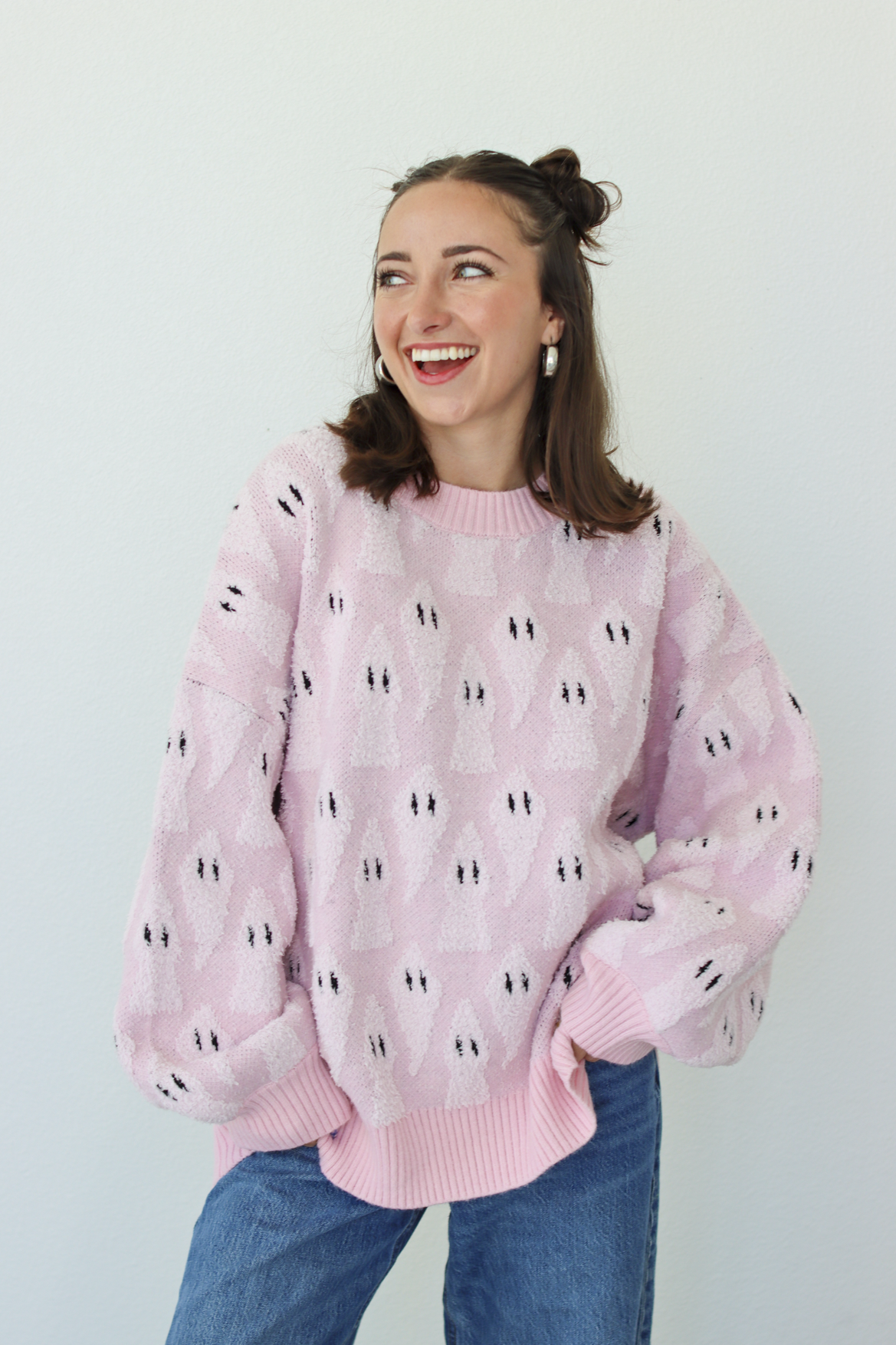 A smiling woman with her hair in two buns wears a light pink fuzzy knit sweater with a repeating black and white ghost pattern, paired with blue jeans, against a plain light gray background.
